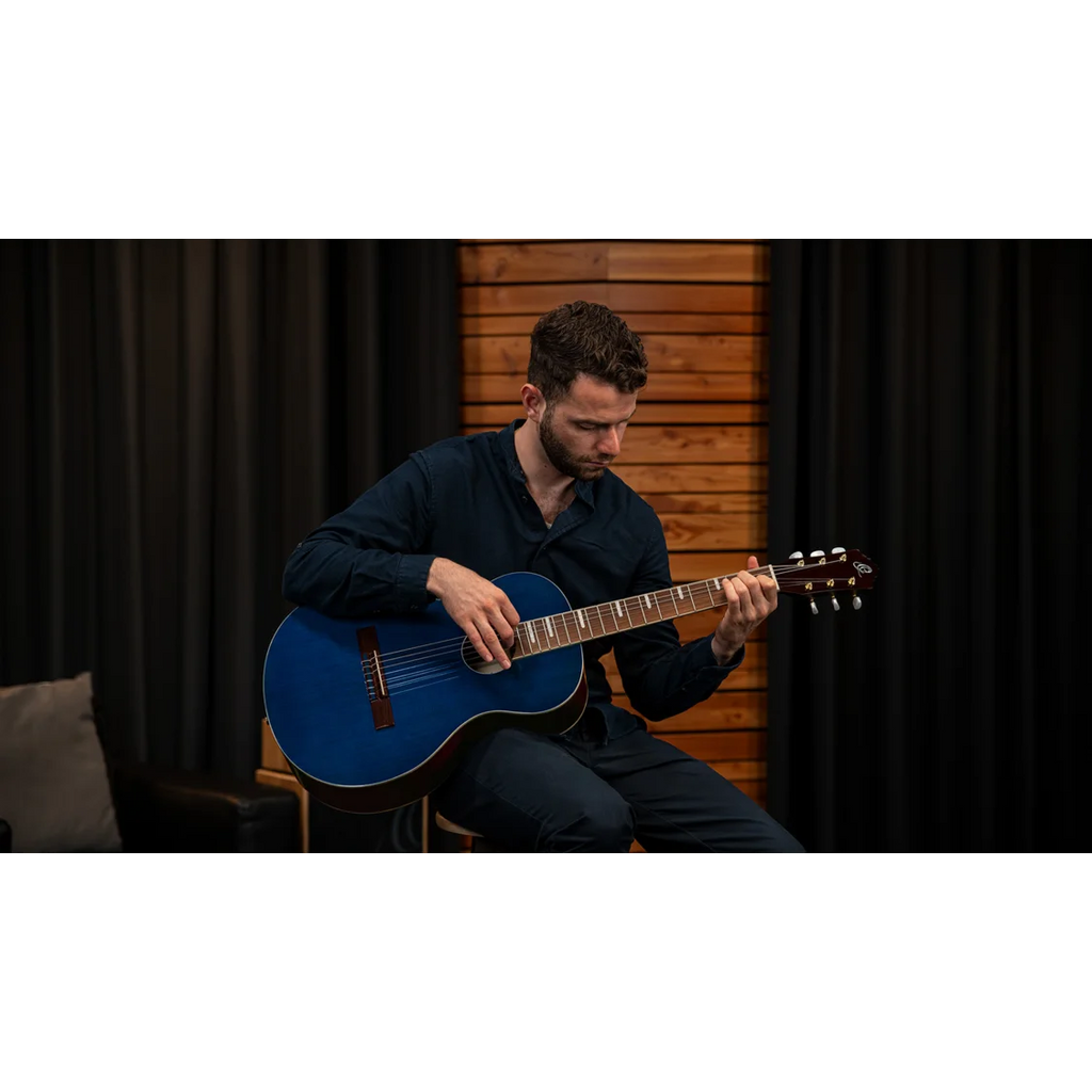Man playing a blue nylon string classical guitar by Ortega in a room with dark curtains and wooden paneling.